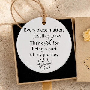 Könnte beinhalten: Ein weißes, rundes Keramikornament mit einer Jutekordel in einer quadratischen Pappschachtel. Das Ornament ist mit schwarzem Text bedruckt, der Every piece matters just like you Thank you for being a part of my journey lautet, sowie einem Puzzleteil-Symbol.