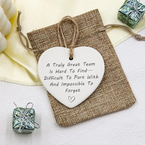 May include: A white heart-shaped ceramic ornament with a jute string for hanging. The ornament has the text "A Truly Great Team Is Hard To Find... Difficult To Part With And Impossible To Forget" and a heart symbol. It rests on a burlap bag.