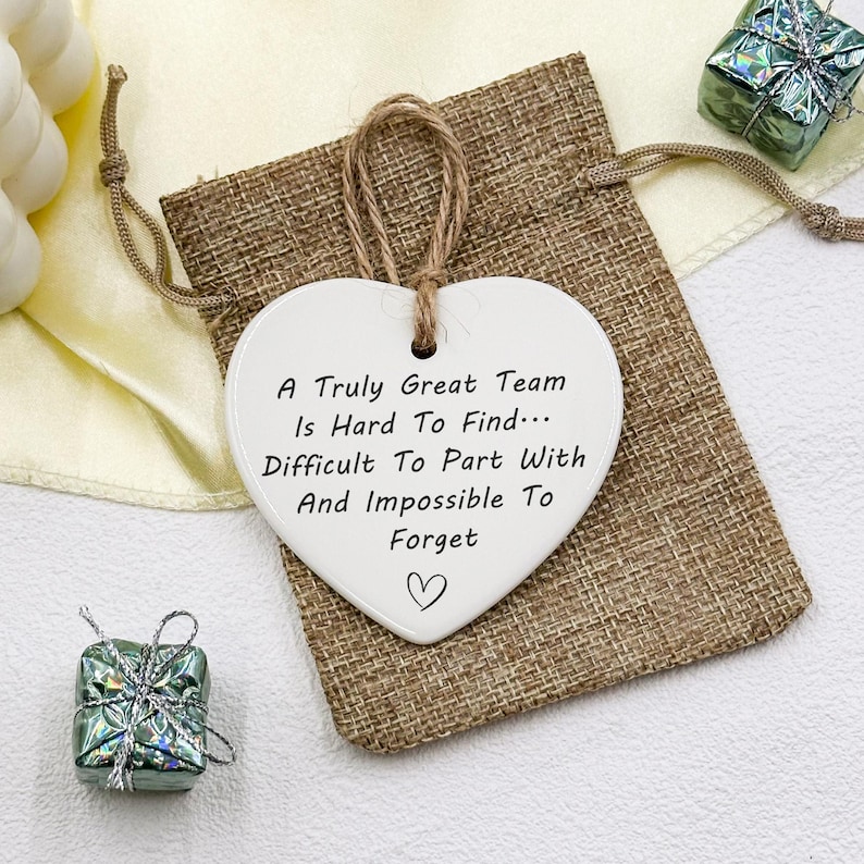 May include: A white heart-shaped ceramic ornament with a jute string for hanging. The ornament has the text "A Truly Great Team Is Hard To Find... Difficult To Part With And Impossible To Forget" and a heart symbol. It rests on a burlap bag.
