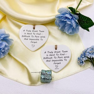 May include: Two heart-shaped ceramic plaques with the text "A Truly Great Team Is Hard To Find... Difficult To Part With And Impossible To Forget" and a heart symbol. The plaques are white with a brown string for hanging. A small gift and blue flowers are also in the image.
