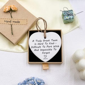 May include: A white heart-shaped ornament with a brown string, displayed in a black box. The ornament has the text "A Truly Great Team Is Hard To Find... Difficult To Part With And Impossible To Forget" and the name "Jessica". A small gift box and a handmade gift box are also visible.