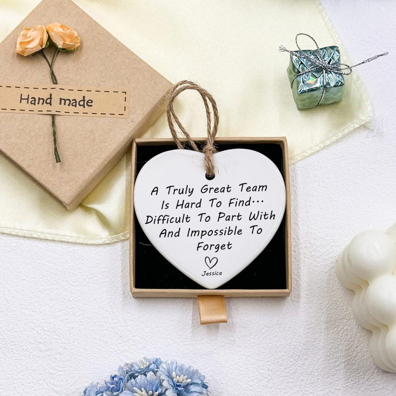 May include: A white heart-shaped ornament with a brown string, displayed in a black box. The ornament has the text "A Truly Great Team Is Hard To Find... Difficult To Part With And Impossible To Forget" and the name "Jessica". A small gift box and a handmade gift box are also visible.