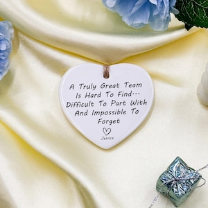 May include: A white heart-shaped ceramic ornament with a jute string for hanging. The ornament has the text "A Truly Great Team Is Hard To Find... Difficult To Part With And Impossible To Forget" and a heart symbol, with the name "Jessica" below.