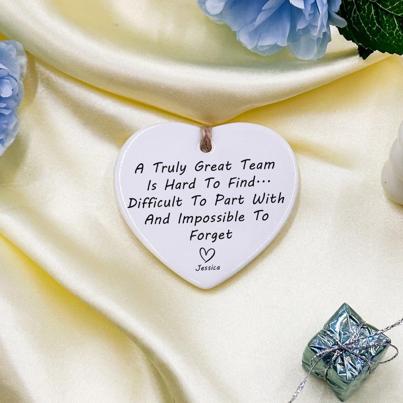 May include: A white heart-shaped ceramic ornament with a jute string for hanging. The ornament has the text "A Truly Great Team Is Hard To Find... Difficult To Part With And Impossible To Forget" and a heart symbol, with the name "Jessica" below.