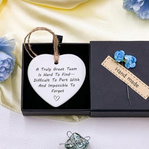 May include: A white heart-shaped ornament with a jute string for hanging, inscribed with the text "A Truly Great Team Is Hard To Find... Difficult To Part With And Impossible To Forget." It rests in a black gift box, with a "Hand made" tag and blue floral accents.
