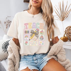 Pode incluir: Uma t-shirt de cor creme com a palavra TEACH em letras grandes e estampadas. Abaixo, um lápis central com um laço de padrão leopardo está rodeado por pequenos gráficos quadrados que incluem lápis de cor, um arco-íris, uma flor, livros, uma mochila, estrelas e o alfabeto.