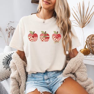 Op de afbeelding: Een crèmekleurig T-shirt met korte mouwen en een afbeelding van drie horizontaal geplaatste appels. Elke appel heeft een patchworkontwerp in rood-, wit- en groentinten met diverse patronen. Onder de derde appel staat de tekst "sweetest".