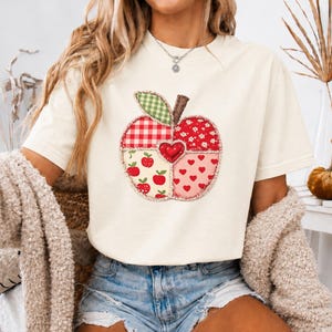 Może przedstawiać: Kremowy t-shirt z krótkim rękawem z dużą, centralną grafiką jabłka. Jabłko składa się z różnych wzorów tkanin w stylu patchwork, w tym czerwonej kratki vichy, małych czerwonych jabłek, białych kwiatów i czerwonych serc z centralnym sercem.