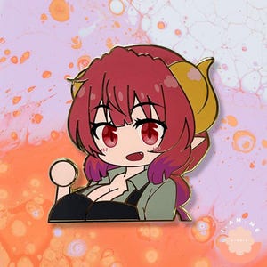 Puede incluir: Un colorido pin de esmalte con un personaje de estilo anime con cabello rojo, ojos rojos y cuernos dorados. El personaje lleva una camisa verde y un top negro. El pin tiene un contorno dorado y un objeto redondo y claro en la mano del personaje.