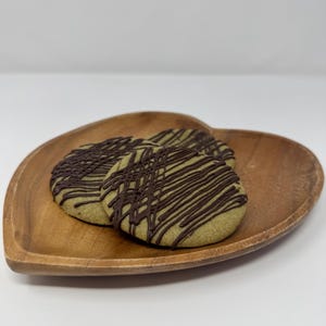 May include: Three round green cookies drizzled with dark chocolate on a heart-shaped wooden plate. The cookies have a light green hue and are arranged in a slightly overlapping pattern, creating a visually appealing presentation.