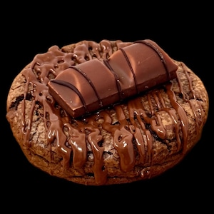 May include: A close-up shot of a chocolate cookie with chocolate drizzle and a chocolate bar on top. The cookie is a dark brown color, and the drizzle is a glossy, rich brown. The chocolate bar is rectangular and has a segmented design.