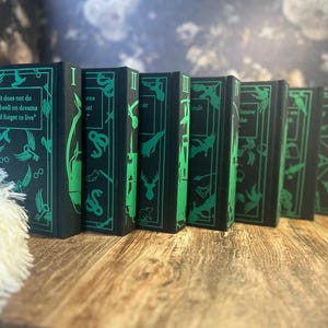 May include: A row of black books with green decorative elements and text. The books feature green illustrations of birds, plants, and other symbols. One book displays the quote, "It does not do to dwell on dreams and forget to live."