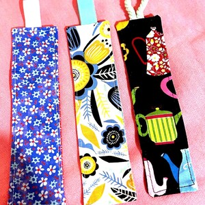 May include: Three fabric bookmarks with different patterns. One has a blue background with white and red flowers, another has a white background with yellow and blue floral designs, and the third has a black background with teapots and other colorful shapes. Each has a ribbon or cord loop.