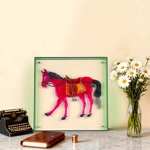 May include: A framed artwork featuring a red horse with a saddle, bridle, and black mane and tail. The horse is set against a cream background within a green-edged frame. A vintage typewriter, leather journal, and flowers are in the foreground.