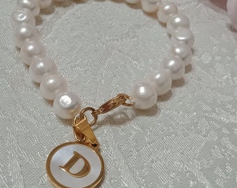 Peut inclure: Un bracelet de perles blanches avec un charme en or représentant la lettre "D" sur fond blanc.