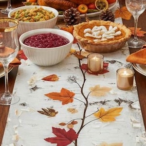 May include: Thanksgiving table setting with a roasted turkey, side dishes, and a decorative runner. The table is set with gold-rimmed glasses, plates, and napkins. A pie and candles add to the festive atmosphere. The runner features a birch bark design with autumn leaves.