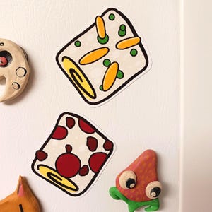 May include: Two square fridge magnets with white backgrounds and black outlines. One features orange and green shapes, the other has red circles. Other magnets in the image include a crescent moon and a strawberry.