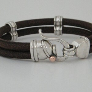 K&ouml;nnte beinhalten: Ein braunes Lederarmband mit einem silbernen Verschluss und einem kleinen ros&eacute;goldenen Akzent. Das Armband ist doppelschichtig und hat ein schlichtes Design.
