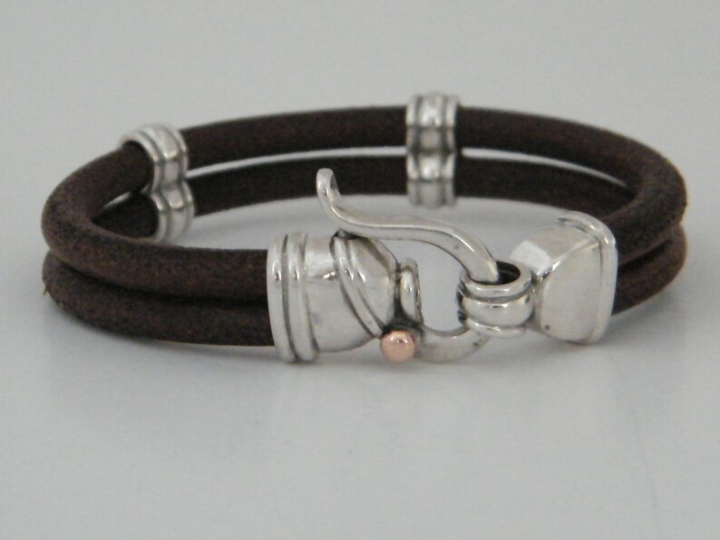 K&ouml;nnte beinhalten: Ein braunes Lederarmband mit einem silbernen Verschluss und einem kleinen ros&eacute;goldenen Akzent. Das Armband ist doppelschichtig und hat ein schlichtes Design.