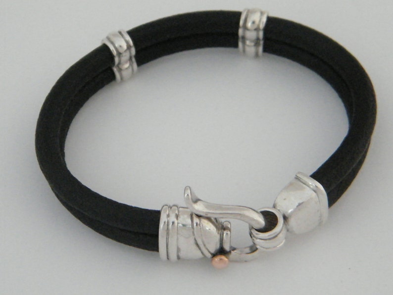 K&ouml;nnte beinhalten: Ein schwarzes Lederarmband mit silbernen Akzenten und einem silbernen Verschluss. Das Armband hat ein schlichtes Design und ist ein klassisches Schmuckst&uuml;ck.