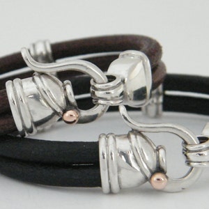 K&ouml;nnte beinhalten: Zweifarbiges Lederarmband mit silbernen Verschl&uuml;ssen und ros&eacute;goldenen Akzenten. Das Armband besteht aus zwei Lederstr&auml;ngen, einem braunen und einem schwarzen.