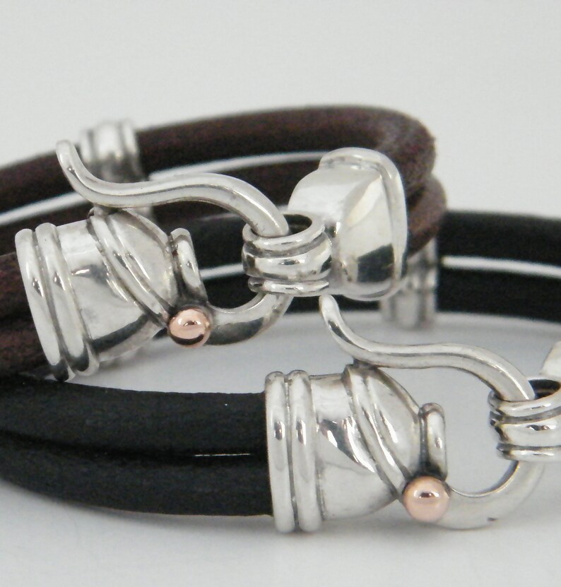 K&ouml;nnte beinhalten: Zweifarbiges Lederarmband mit silbernen Verschl&uuml;ssen und ros&eacute;goldenen Akzenten. Das Armband besteht aus zwei Lederstr&auml;ngen, einem braunen und einem schwarzen.