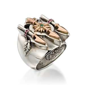 May include: A silver ring with gold and ruby accents. The ring features a central design with a green gemstone and gold arrow-like elements. Additional gold and ruby details adorn the ring's surface, creating a unique and ornate appearance.