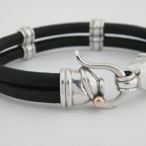 K&ouml;nnte beinhalten: Ein schwarzes Lederarmband mit einem silbernen Verschluss und einem kleinen Akzent aus Ros&eacute;gold. Das Armband ist doppelschichtig und hat eine strukturierte Oberfl&auml;che.