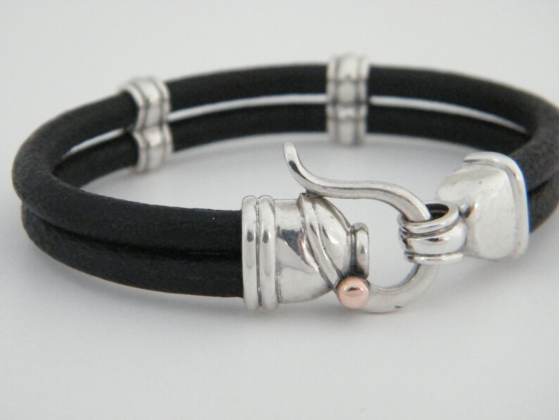 K&ouml;nnte beinhalten: Ein schwarzes Lederarmband mit einem silbernen Verschluss und einem kleinen Akzent aus Ros&eacute;gold. Das Armband ist doppelschichtig und hat eine strukturierte Oberfl&auml;che.