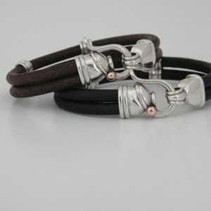 K&ouml;nnte beinhalten: Zwei Lederarmb&auml;nder mit silbernen und goldenen Verschl&uuml;ssen. Die Armb&auml;nder sind braun und schwarz.