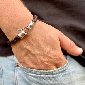 Puede incluir: Un brazalete de cuero marrón con un cierre plateado. El brazalete está trenzado y tiene un cierre de gancho.