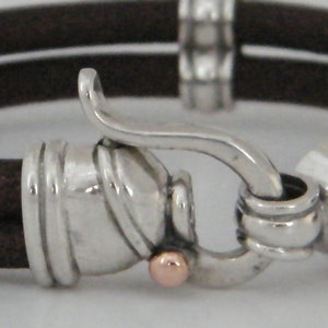 K&ouml;nnte beinhalten: Ein braunes Lederarmband mit einem silbernen Verschluss und einem ros&eacute;goldenen Akzent. Der Verschluss ist ein Haken- und &Ouml;sen-Design.