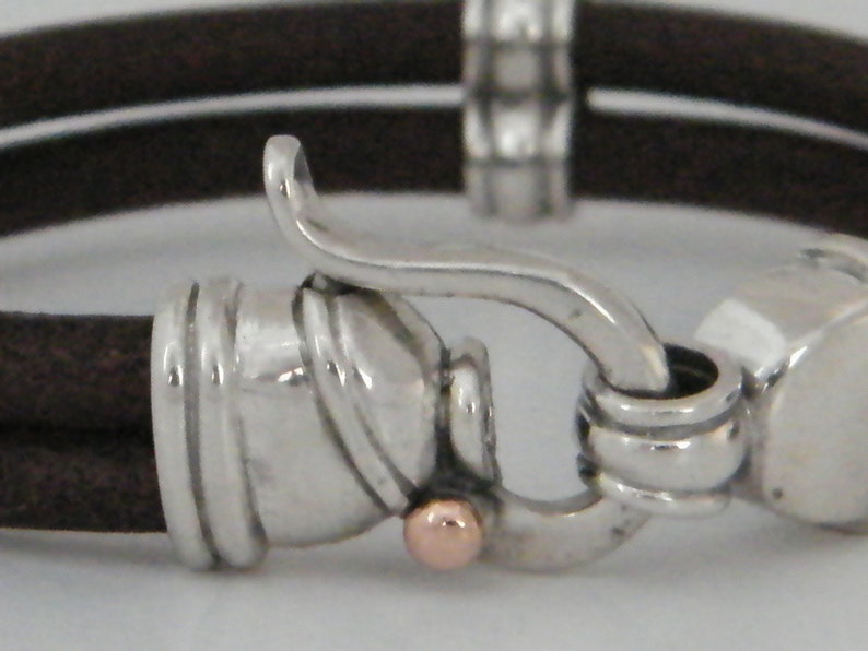 K&ouml;nnte beinhalten: Ein braunes Lederarmband mit einem silbernen Verschluss und einem ros&eacute;goldenen Akzent. Der Verschluss ist ein Haken- und &Ouml;sen-Design.
