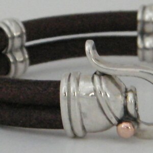 K&ouml;nnte beinhalten: Ein braunes Lederarmband mit einem silbernen Verschluss und einem kleinen goldenen Akzent. Das Armband ist doppelschichtig und hat ein schlichtes Design.