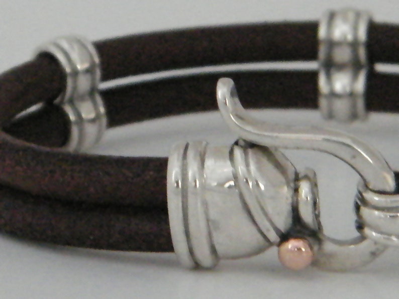K&ouml;nnte beinhalten: Ein braunes Lederarmband mit einem silbernen Verschluss und einem kleinen goldenen Akzent. Das Armband ist doppelschichtig und hat ein schlichtes Design.