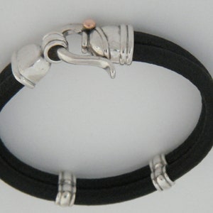 K&ouml;nnte beinhalten: Ein schwarzes Lederarmband mit silbernen und ros&eacute;goldenen Akzenten. Das Armband hat einen Hakenverschluss und drei silberne Perlen.