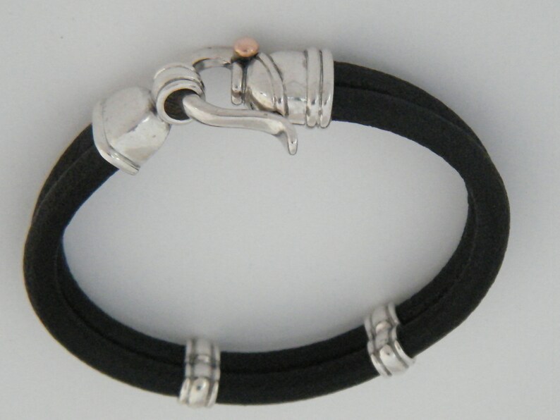 K&ouml;nnte beinhalten: Ein schwarzes Lederarmband mit silbernen und ros&eacute;goldenen Akzenten. Das Armband hat einen Hakenverschluss und drei silberne Perlen.