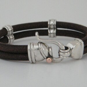 K&ouml;nnte beinhalten: Ein braunes Lederarmband mit einem silbernen Verschluss und einem kleinen ros&eacute;goldenen Akzent. Das Armband ist doppelschichtig und hat ein schlichtes Design.