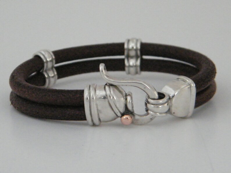 K&ouml;nnte beinhalten: Ein braunes Lederarmband mit einem silbernen Verschluss und einem kleinen ros&eacute;goldenen Akzent. Das Armband ist doppelschichtig und hat ein schlichtes Design.