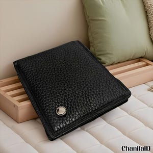Peut inclure: Un portefeuille en cuir noir avec une surface texturée et une fermeture à bouton argenté. Le portefeuille de forme carrée semble fermé. En arrière-plan, un plateau en bois et un coussin vert.