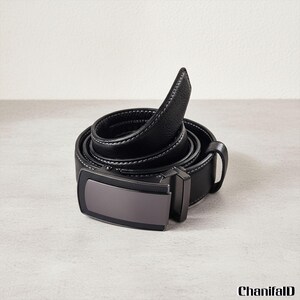 Puede incluir: Un cinturón de cuero negro con hebilla de metal pavonado y costuras blancas. El cinturón está enrollado, mostrando su textura y diseño. La hebilla es rectangular y tiene un acabado mate.