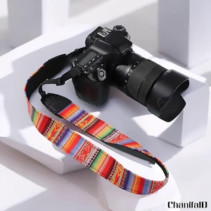 May include: A black DSLR camera with a lens and a colorful patterned camera strap. The strap features a vibrant design with red, yellow, green, and blue geometric patterns. The camera is resting on a white surface.