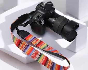 Könnte beinhalten: Eine schwarze DSLR-Kamera mit Objektiv und einem farbenfrohen Kameragurt. Der Gurt hat ein lebendiges Design mit roten, gelben, grünen und blauen geometrischen Mustern. Die Kamera liegt auf einer weißen Oberfläche.