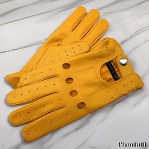 May include: A pair of bright yellow driving gloves made of leather. The gloves have a classic design with finger holes and circular perforations for ventilation. A silver snap closure is visible on the wrist. The gloves are displayed on a marble surface.