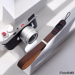 Puede incluir: Una cámara Leica plateada y negra con una correa de muñeca marrón. La cámara tiene una lente plateada y detalles en rojo. La correa de muñeca es marrón con un conector negro y rojo y una pieza de ajuste de metal plateado.