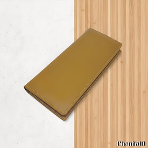 Peut inclure: Un portefeuille en cuir de couleur moutarde, de forme rectangulaire, avec une bordure cousue. Le portefeuille est présenté sur un fond divisé en blanc moucheté et en bois clair.