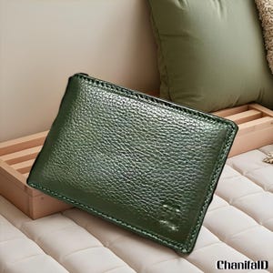 Puede incluir: Una cartera de cuero verde oscuro con una superficie texturizada. La cartera es rectangular y parece estar cerrada. Las costuras son visibles alrededor de los bordes. La cartera está sobre una superficie blanca.