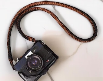Könnte beinhalten: Schwarze und blaue Vintage-Kamera mit einem braunen und schwarzen Seilriemen. Die Kamera hat ein silbernes Objektiv und eine schwarze Ledertasche. Der Riemen besteht aus gewebtem Material. Die Kamera liegt auf einer weißen Marmoroberfläche.