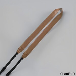 May include: A tan leather camera strap with white stitching and black nylon straps. The camera strap has metal rings for attachment. The strap is angled across a light grey background.