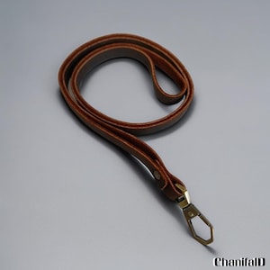 May include: A brown leather lanyard with a metal clasp. The lanyard is made of a single strip of leather, looped and stitched together. The metal clasp is a bronze color and is attached to the end of the lanyard.